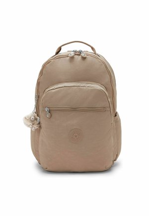 Beiger Rucksack mit zwei Reißverschlussfachern, seitlichen Taschen, einem runden Logo auf der Vorderseite und einem kleinen Plüsch-Schlüsselanhänger am Reißverschluss.