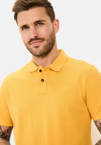 Polo t-shirt in felgeel, korte mouwen, textuurstof, drie-knopen sluiting met zwarte knopen, en een klein logo op de borst.