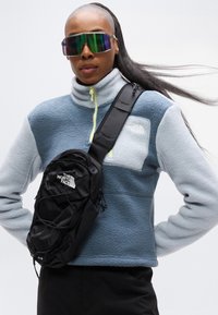 Φούτερ από φλις σε ανοιχτό μπλε και γκρι με ψηλό γιακά, σε συνδυασμό με μια μαύρη τσάντα ώμου που διαθέτει λογότυπο και ελαστικό κορδόνι μπροστά.