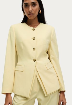 Blazer giallo dal design strutturato, con scollatura a V, cinque bottoni arrotondati e due tasche frontali. Tessuto liscio e sartoriale, maniche lunghe.