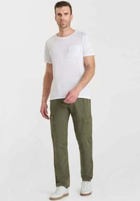 Man die staat en een wit T-shirt met korte mouwen, olijfgroene cargobroek en witte sneakers draagt tegen een effen lichte achtergrond.
