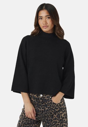 Frau mit mittellangem dunklem Haar, die einen schwarzen Langarmpullover und eine Hose mit Leopardenmuster trägt, steht mit einer Hand in der Tasche.