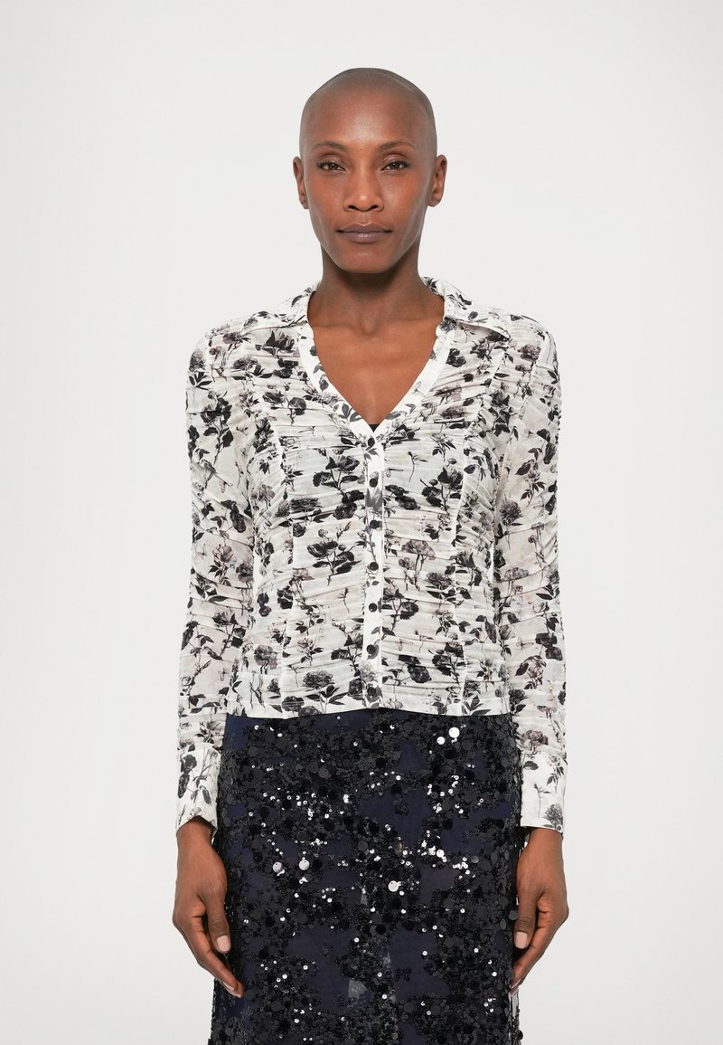 Short-sleeve blouse in white with black floral print, featuring a V-neck, button closure, and textured fabric. Worn with a sequined dark skirt.
