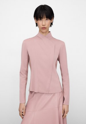 Femme aux cheveux courts noirs portant un blazer rose clair cintré et une jupe assortie, debout devant un fond blanc uni.