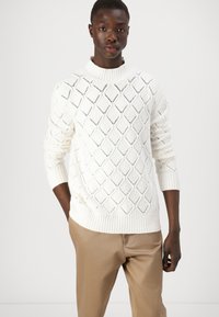 Maglione bianco lavorato a maglia con motivo a diamante, polsini e orlo a costine, abbinato a pantaloni beige. Texture morbida e design a vestibilità ampia.