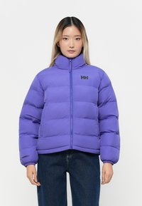 Giacca puffo viola con colletto alto, chiusura con zip e maniche voluminose. Presenta un logo sul petto e polsini elasticizzati. Indossata sopra jeans scuri.