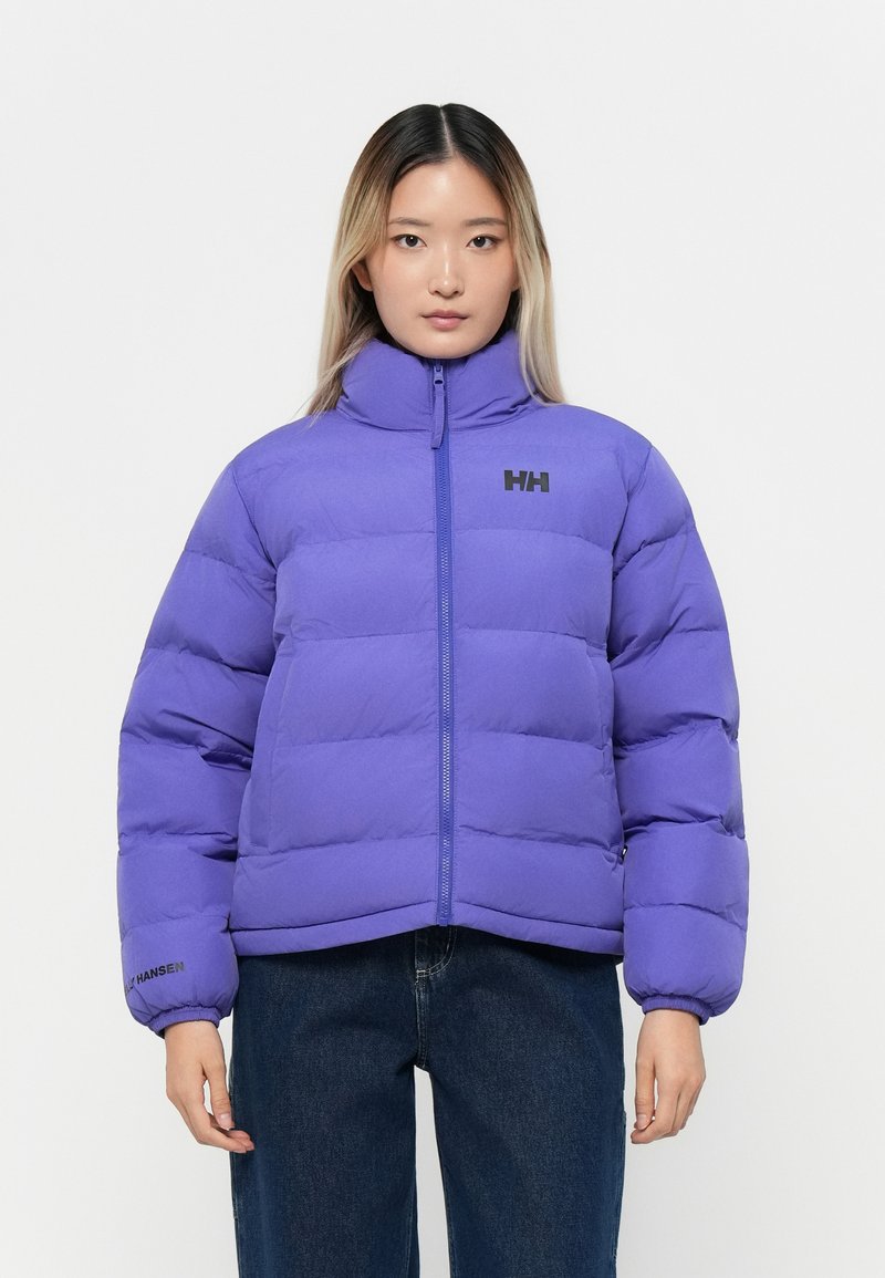 Veste matelassée violette avec un col haut, une fermeture éclair et des manches bouffantes. Présente un logo sur la poitrine et des poignets élastiques. Portée par-dessus un jean foncé.
