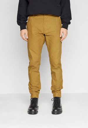 Pantaloni tapered beige con quattro tasche, realizzati in un tessuto resistente; indossati con stivali neri stringati; presentano una texture liscia e un design essenziale.