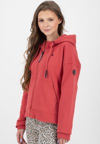 Felpa rossa con zip, realizzata in materiale morbido, dotata di due tasche frontali, cordini regolabili e una toppa con logo sulla manica.