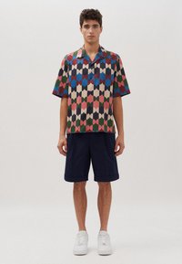 Kurzärmliges Hemd mit Button-Down-Kragen in buntem geometrischen Muster mit Nuancen von Rot, Grün, Blau und Beige, kombiniert mit marineblauen Shorts und weißen Sneakers.