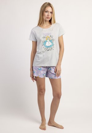 Mujer de pie descalza con camiseta gris y pantalones cortos morados con estampados de Alicia en el País de las Maravillas sobre un fondo claro liso.