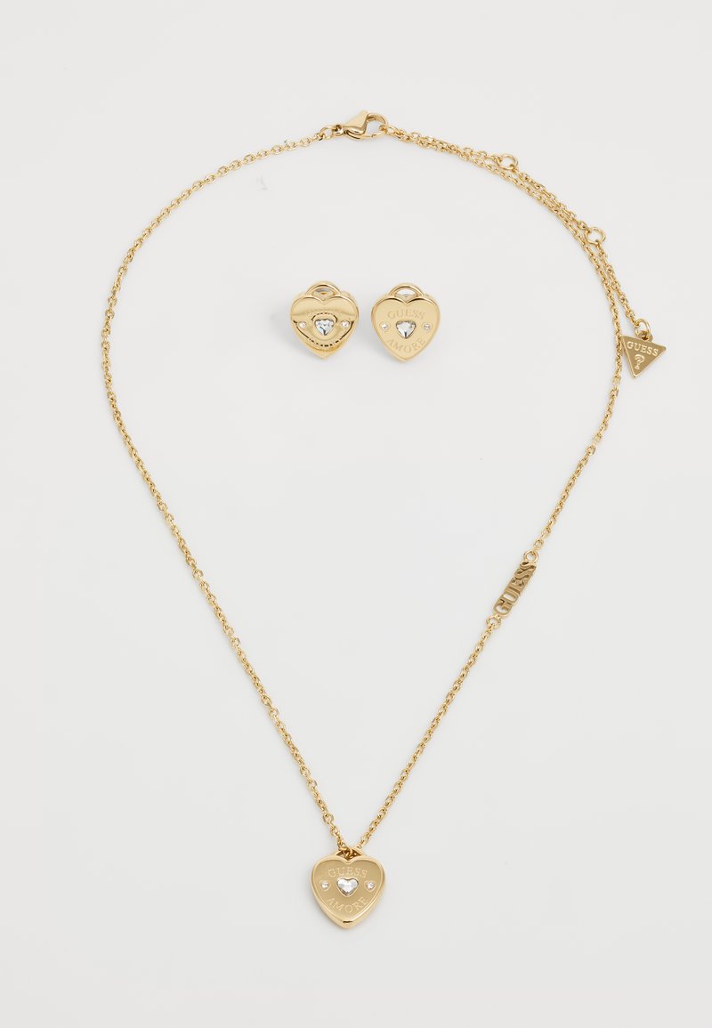 Gold necklace with heart-shaped pendant, featuring a small clear gemstone and "GUESS AMORE" engraving. Includes matching heart-shaped earrings.