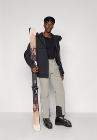 Costum de schi care include o jachetă neagră cu buzunare, pantaloni gri deschis cu accente negre și bocanci de schi negri. Ține în mână schiuri cu grafică roz.