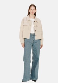 Kvinne iført en beige knappet jakke med store lommer, hvit topp, vide blå jeans og svarte spisse sko, stående mot hvit bakgrunn.