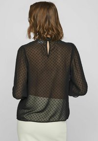 Sort gennemsigtig bluse med prikmønster, der har en nøglehul-udskæring bagpå, lange puffærmer og en glat kant. Designet med et lagdelt udseende.