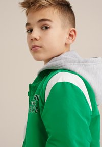 Zelena bomber jakna siva podstava sa kapuljačom, rebrasti ovratnik, bijeli akcenti na ramenima i izvezeni natpis, sa glatkom, sjajnom površinom.