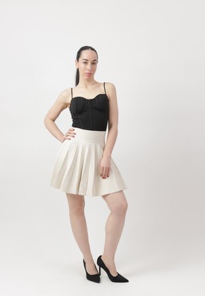 Mujer posando con la mano en la cadera, vistiendo un top negro sin mangas, falda blanca plisada y tacones altos negros contra un fondo blanco liso.