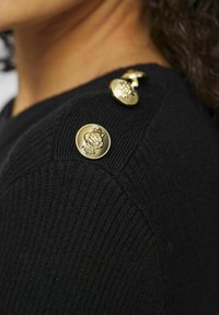Svart stickad tröja med tre gyllene knappar med intrikata lejonmotiv på axeln, som har en ribbad textur och skräddarsydd passform.