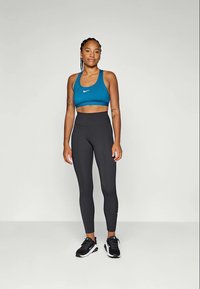 Donna in reggiseno sportivo blu, leggings neri a vita alta e sneaker neri, in piedi contro uno sfondo chiaro e uniforme.