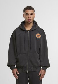 Szürke, cipzáros kapucnis pulóver, pamut keverékből készült, elülső zsebbel, rojtos alsó résszel és kerek, narancssárga logóval a mellrészen.