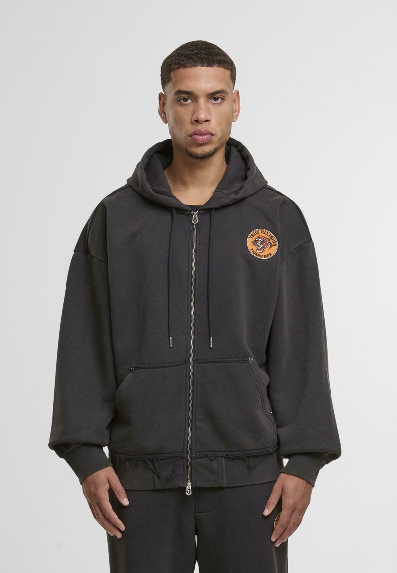 Szürke, cipzáros kapucnis pulóver, pamut keverékből készült, elülső zsebbel, rojtos alsó résszel és kerek, narancssárga logóval a mellrészen.