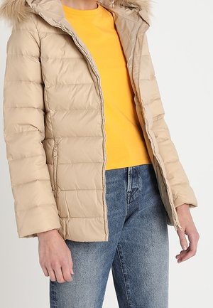 Mujer con chaqueta beige acolchada con capucha con borde de piel, desabrochada sobre una camiseta amarilla y vaqueros azules sobre un fondo liso.