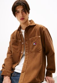 Brązowa koszula w korduroy z dwoma kieszeniami na piersi, białymi guzikami i małą naszywką z logo "TOMMY JEANS" na lewej kieszeni. Nosi się ją na białym t-shircie.