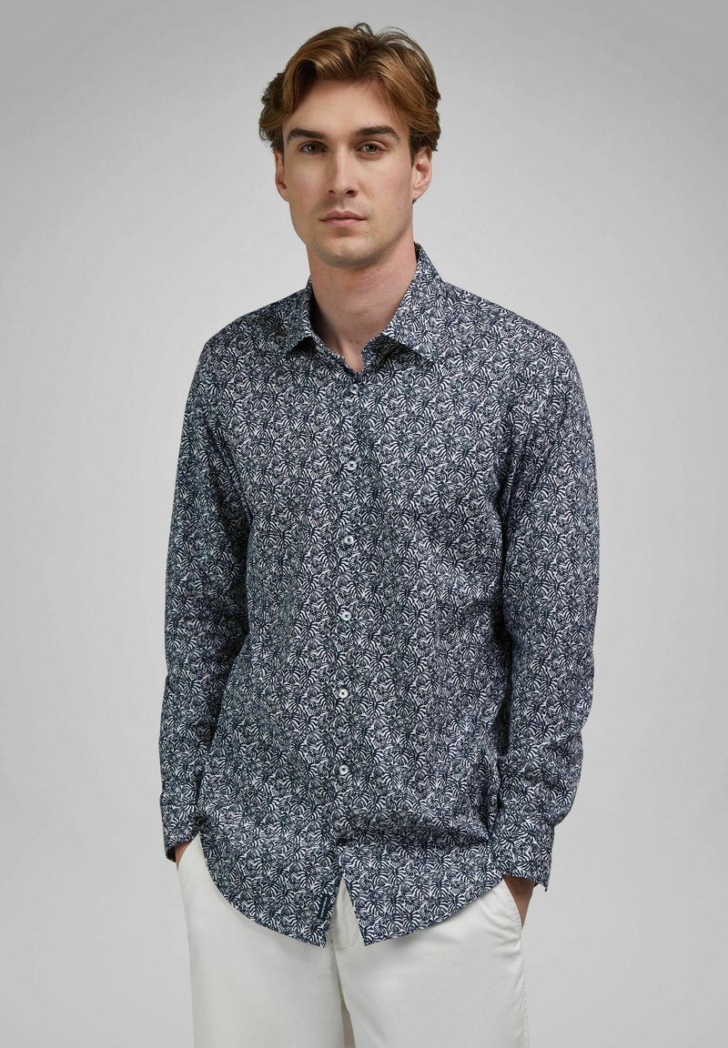 Giovane uomo in piedi con le mani in tasca, indossa una camicia blu scuro con motivo a bottoni e pantaloni bianchi su uno sfondo grigio semplice.