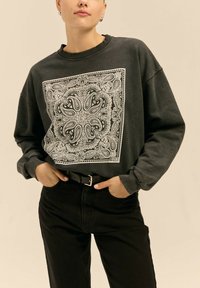 Przedłużony czarny sweatshirt z białym wzorem paisley; ściągaczowy dekolt i mankiety; zestawiony z czarnymi spodniami na wysokim stanie i czarnym paskiem.