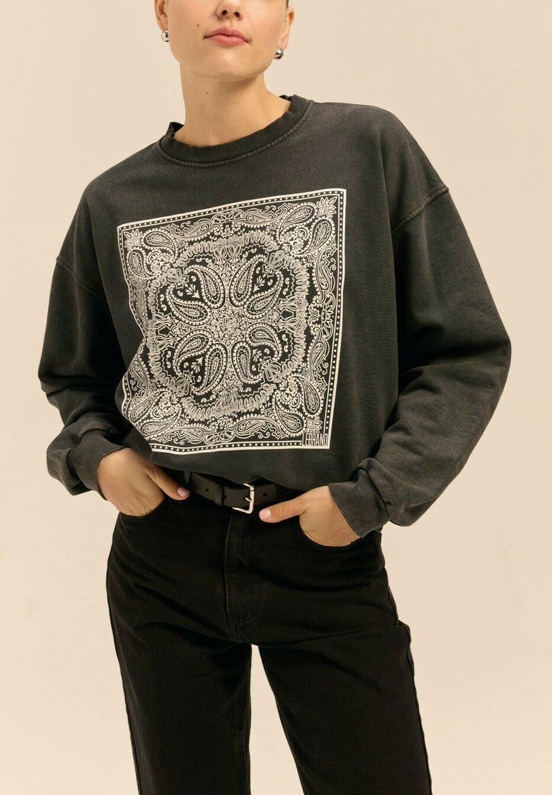 Przedłużony czarny sweatshirt z białym wzorem paisley; ściągaczowy dekolt i mankiety; zestawiony z czarnymi spodniami na wysokim stanie i czarnym paskiem.