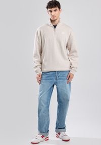 Pullover beige chiaro con colletto a mezza zip, abbinato a jeans azzurri a vestibilità morbida e sneakers bianche con dettagli rossi. Tessuto di consistenza liscia.