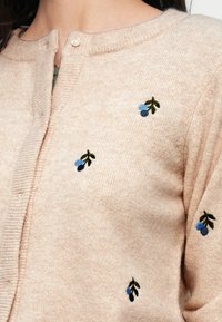 Ροζ ανοιχτή ζακέτα με στρογγυλή λαιμόκοψη, που διαθέτει κεντητά σχέδια με μπλε και πράσινα μούρα και λευκά κουμπιά κατά μήκος του μπροστινού μέρους.