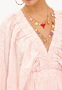 Roze, geborduurde jurk met een gerimpelde halslijn en mouwen. Gelaagde gouden kettingen met kleurrijke bedels, waaronder zeethema-ontwerpen.