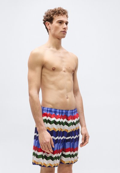 Νεαρός άνδρας με κατσαρά μαλλιά που φοράει πολύχρωμα σορτς με γεωμετρικά σχέδια, στέκεται μπροστά σε απλό ανοιχτό γκρι φόντο και κοιτάζει πλάγια.