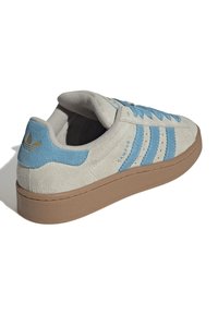 Sneakers in suede grigio con accenti blu, tre strisce e una suola in gomma marrone dall'aspetto testurizzato. Dettaglio del logo dorato sul tallone.