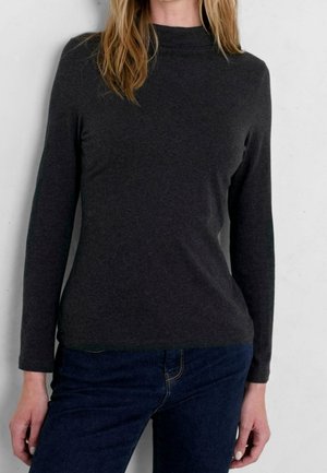 Femme portant un haut moulant à manches longues gris foncé et un jean bleu foncé, debout contre un mur gris clair.