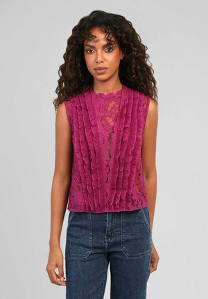 Femme aux cheveux bouclés portant un haut en dentelle magenta sans manches avec des panneaux froncés à l'avant et un jean bleu taille haute sur un fond uni.