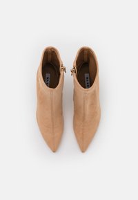 Stivali ankle in finta suede beige con punta affusolata, cerniere laterali e dettagli cuciti. Tessitura liscia con un design snello e aderente.