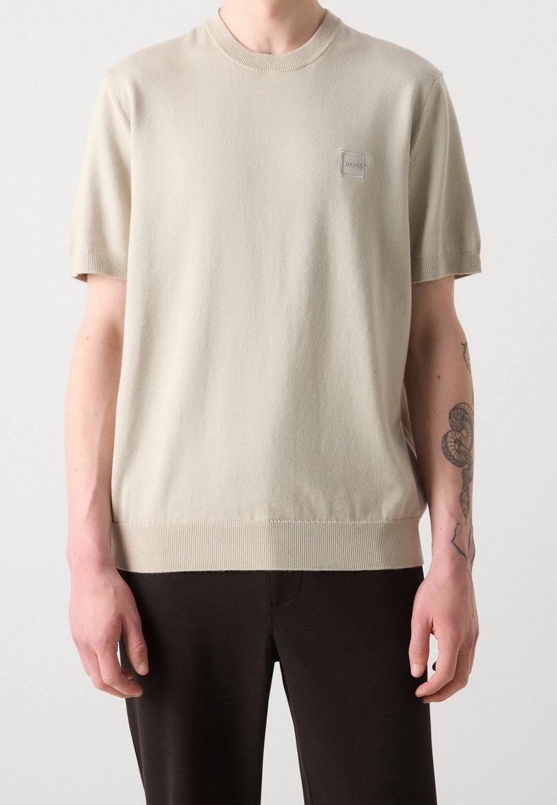 Homme portant un t-shirt beige à manches courtes avec col rond, bords côtelés et petit logo poitrine, associé à un pantalon noir et un tatouage visible sur le bras.