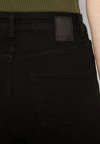 Μαύρα τζιν με λείο φινίρισμα, με ένα ορθογώνιο δερμάτινο λογότυπο με την ένδειξη "PIECES" στη μέση και λεπτομέρεια στην πίσω τσέπη.