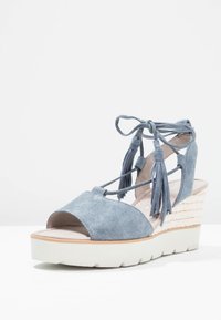 Blue suede wedge sandal with an open toe, lace-up design, and decorative tassels; features a textured woven platform and white rubber sole.
