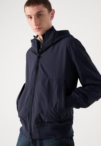 Navyblauwe hoodie gemaakt van lichtgewicht materiaal, met een rits aan de voorkant, twee zijzakken en elastische manchetten.
