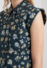 Blusa floreale verde scuro con maniche corte, colletto e molteplici motivi di fiori bianchi e gialli, con bottoni marroni.