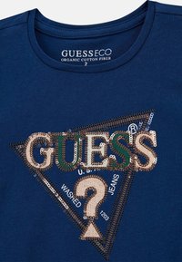 Tamsiai mėlynas organinės medvilnės marškinėliai su dekoratyvine trikampio formos blizgučiais logotipu, kuriame užrašyta "GUESS" auksine ir žalia spalvomis, bei dekoratyvinis tekstas žemiau.