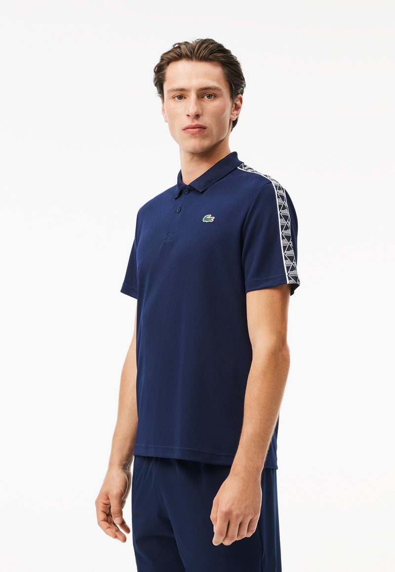 Marineblaues Poloshirt mit kurzen Ärmeln, einem Kragen und einem grünen Logo. Es verfügt über einen kontrastierenden geometrischen Musterbesatz entlang der Schultern.