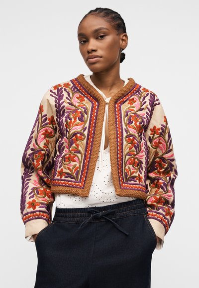 Femme aux cheveux tressés portant une veste courte beige avec une broderie florale colorée, un chemisier blanc et un pantalon sombre à cordon de serrage.