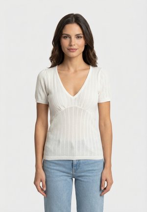 Vrouw met donker haar, gekleed in een wit kortermig gebreid shirt met V-hals en lichtblauwe spijkerbroek, staand tegen een effen achtergrond.