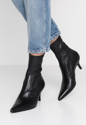 Bottes noires à cheville avec des bouts pointus, en cuir lisse. Présentent une tige ajustée et un talon mince et incliné. Portées avec un jean en denim retroussé.