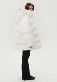 Biała kurtka puchowa z wysokim kołnierzem i kapturem, z pikowanym wzorem i błyszczącym materiałem, w połączeniu z czarnymi, prążkowanymi spodniami i spiczastymi butami.