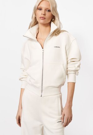 Blondynka w kremowej kurtce zapinanej na zamek oraz dopasowanych spodniach z logo "LAMANIA" na klatce piersiowej i udzie, stojąca na białym tle.